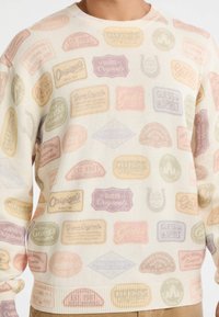 Witte gebreide trui met een patroon van kleurrijke, vintage-stijl merklabels in pastelkleuren, voorzien van een ronde hals en een geribbelde zoom.