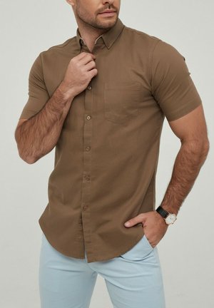 Chemise marron à manches courtes avec un col, dotée d'une poche poitrine et d'une texture douce, associée à un pantalon bleu clair.