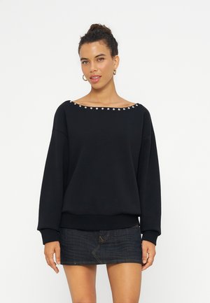 Femme aux cheveux bouclés portant un haut noir à manches longues avec des détails en perles au niveau du col et une mini-jupe en jean foncé sur fond blanc.