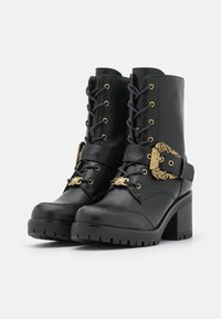 Stivali neri in pelle fino alla caviglia con tacchi chunky, lacci frontali, suole spesse e dettagli decorativi in oro con fibbie e accenti metallici.