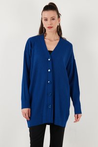 Žena oblečená v modrom dlhom rebrovanom kardigáne s veľkými gombíkmi, čiernych nohaviciach, zlatých dlhých náušniciach a zlatom reťazovom náhrdelníku na bielom pozadí.