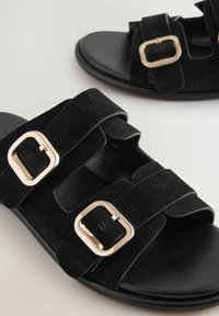 Sandales en daim noir avec deux sangles réglables ornées de boucles en métal doré et une semelle plate lisse noire. Le matériau texturé apporte de la profondeur.