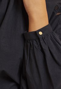 Blusa di cotone blu navy con design delle maniche arricciate, caratterizzata da un accento di bottone dorato al polsino. Tessuto morbido e traspirante.