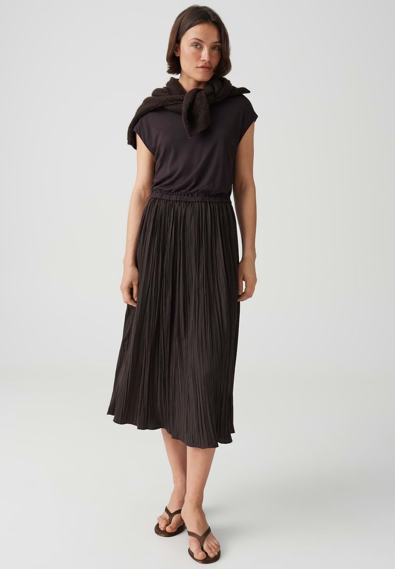 Woman wearing a black sleeveless top, long pleated black skirt, sandals, and a sweater tied around her shoulders against a plain background.