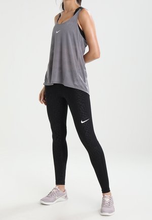 Donna che indossa un top grigio senza maniche, leggings neri Nike con motivo sottile e scarpe da ginnastica Nike rosa chiaro, in piedi con le mani dietro la schiena.