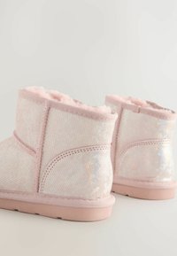 Stivali alla caviglia foderati di peluche rosa con una finitura glitterata sottile, suole rotonde e cuciture visibili su uno sfondo chiaro e semplice.