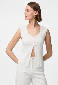 Witte geribbelde mouwloze top met een gekartelde halslijn, voorzien van een vooropening en kleine knopen. Gemaakt in combinatie met witte broek.