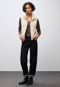 Krémová vesta z umělé kožešiny se zipem vpředu přes hnědý svetr s výstřihem do V, doplněná o tmavé džíny s širokými nohavicemi a černé kotníkové boty.