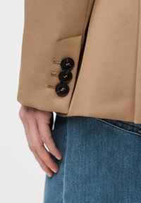 Μπεζ σακάκι με υφή υφάσματος, διακοσμημένο με τέσσερα μαύρα κουμπιά στο μανίκι και εφαρμοστή γραμμή. Στο κάτω μέρος φαίνονται τζιν παντελόνια.
