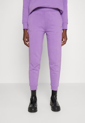 Persoon die lila joggingbroek en bijpassend sweatshirt draagt, gecombineerd met zwarte chunky enkellaarsjes, staand tegen een effen lichte achtergrond.