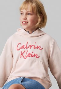 Felpa rosa con tasca frontale, presenta la scritta in rilievo "Calvin Klein" in rosso. Realizzata in tessuto morbido, con cappuccio e polsini a coste.