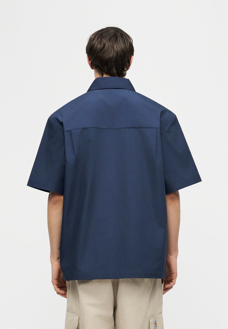 Personne aux cheveux courts et foncés, portant une chemise ample à manches courtes bleu marine et un short cargo beige, vue de dos.
