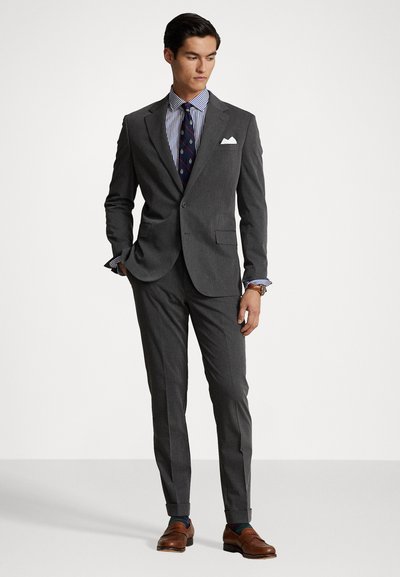 Grijze colbert en broek met notch-lapels, gedragen over een gestreept overhemd en een geblokte stropdas. Bruine leren schoenen maken de outfit compleet.