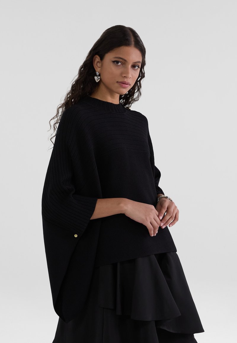 Schwarzer gerippter Strickpullover mit hohem Kragen und weiten Ärmeln, versehen mit einem goldenen Knopfakzent an einem Ärmel. Getragen über einem geschichteten schwarzen Rock.
