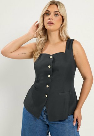 Femme aux longs cheveux blonds portant un haut noir sans manches boutonné et un jean bleu, posant avec une main près de sa tête.