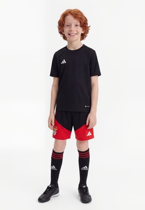 Garçon souriant aux cheveux roux portant un t-shirt de sport noir, un short de football rouge et noir, des chaussettes Adidas noires et des crampons noirs, debout sur un fond blanc.