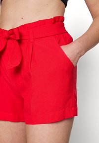Gros plan sur une personne portant un short rouge taille haute avec une ceinture nouée et la main droite placée dans la poche avant.