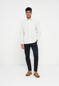 Cămașă albă cu dungi verticale, pantaloni bleumarin și pantofi bej. Modelul stă în fața unui fundal alb, cu mâinile relaxate.
