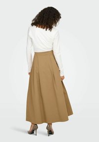 Jupe longue plissée marron en coton, dotée d'une taille haute et d'une coupe fluide ; associée à un haut blanc à manches longues et des talons gris.