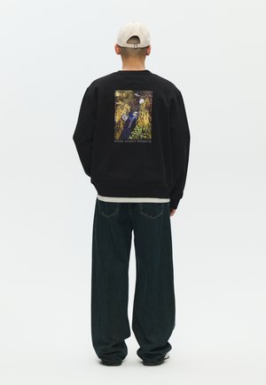 Muškarac u crnoj majici s natpisom i fotografijom prirode na leđima, tamnim širokim trapericama i bež kapa, okrenut leđima prema jednobojnoj pozadini.