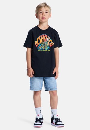 Zwart T-shirt met kleurrijke grafische print, denim shorts en zwart-witte sneakers. Het ontwerp toont levendige motorafbeeldingen.