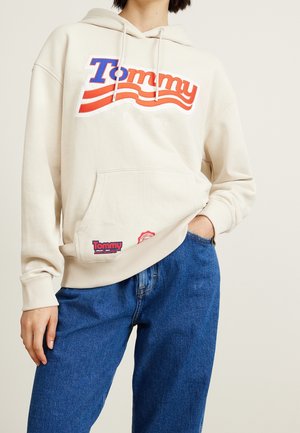 Person trägt cremefarbenen Hoodie mit "Tommy"-Logo in Rot und Blau, kombiniert mit blauen Jeans, Hand in der Tasche, neutraler Hintergrund.