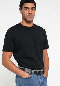 Mustast puuvillast lühikeste varrukatega t-särk ja ümmarguse kaelusega, koos siniste teksade ja musta vööga, esitades lõdvestunud istuvust.