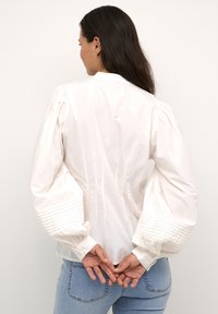 Weiße Bluse mit Puffärmeln und gerippten Bündchen, aus leichtem Baumwollstoff, mit lockerer Passform, getragen mit hellblauen Jeans.