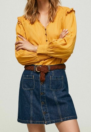 Žena nosící žlutou halenku s dlouhým rukávem a volánky a džínovou sukni s knoflíky vpředu, předními kapsami a hnědým pleteným páskem.