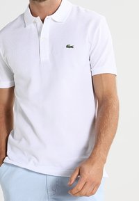 Polo blanc en coton, doté d'une patte de boutonnage à trois boutons et d'un logo crocodile vert sur la poitrine gauche. Manches courtes, coupe classique.