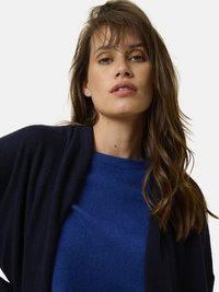 Marinblå cardigan ovanpå en blå tröja, båda i mjukt material. Cardiganen har en öppen design fram och minimal sömnadsdetalj.