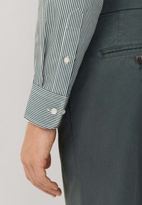 Groene en witte gestreepte blouse met knopen en een contrasterende manchette. Grijze stof zichtbaar, met een knoopdetail. Soepele textuur.