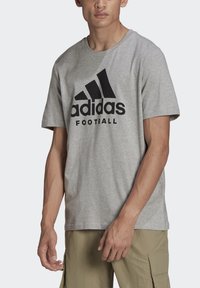 T-shirt grigio a maniche corte in cotone, con un grande logo Adidas nero e la parola "FOOTBALL" sotto di esso. Design semplice e casual.