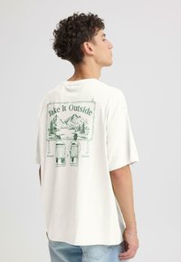 T-shirt en coton blanc à coupe décontractée, avec un motif vert représentant un panneau avec des montagnes et deux chaises dans le dos.