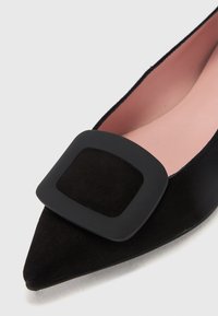 Melna zamšīga kurpe ar izteikta pirksta formu, kurai priekšā ir liela kvadrātveida matēta melna sprādze un mīksta gaiši rozā iekšējā oderējuma.