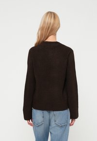 Suéter de malha castanho com mangas longas, gola redonda e bainha canelada. Usado com jeans azul claro, exibindo um design texturizado e aconchegante.