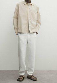 Camicia beige con bottoni e taschino sul petto, abbinata a pantaloni bianchi larghi e sandali pianeggianti marroni su una superficie texturizzata.