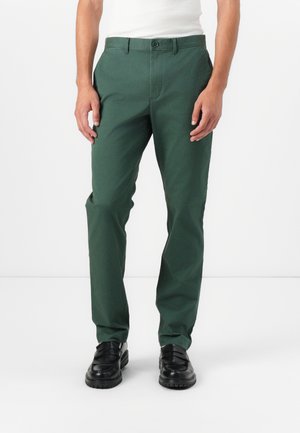 Pantalon en coton vert avec une coupe slim, poches avant et fermeture à un seul bouton. Texture lisse, design plat à l'avant, associé à des mocassins noirs.