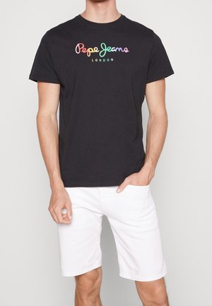 Mężczyzna w czarnej koszulce Pepe Jeans London z tęczowym logo i białych szortach do kolan, stojący na jednolitym jasnym tle.