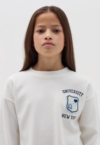 Witte gebreide sweatshirt met een blauw en zwart geborduurd universiteitswapen op de borst, met de tekst "UNIVERSITY" en "NEW YORK".