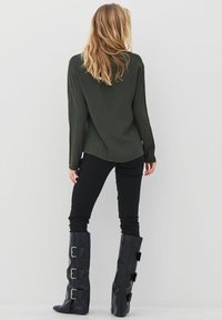 Tamsiai žalia ilgomis rankovėmis palaidinė su kreivėmis kraštais, derinama su juodomis kelnėmis ir juodais aukštakulniais batais iki kelių, turinčiais tris sidabro spaustukus.