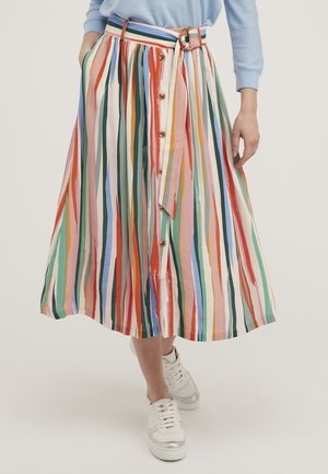 Midi-rok met verticale strepen in verschillende kleuren, voorzien van knopen en een stoffen riem, gecombineerd met een lichtblauwe trui en witte sneakers, staand op een witte vloer.