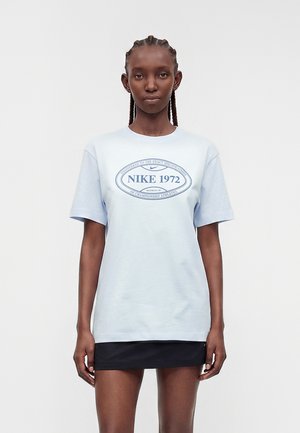 Žena s pletenicama, nosi svijetloplavu Nike 1972 majicu i crnu suknju, stoji ispred jednobojne pozadine, okrenuta prema naprijed.