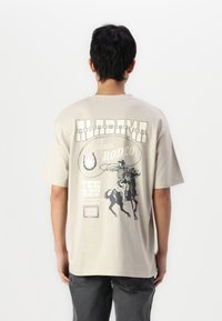 Camiseta de manga corta beige con un estampado gráfico que presenta a un vaquero en un caballo desbocado, el texto "Alabama" y adornos decorativos.