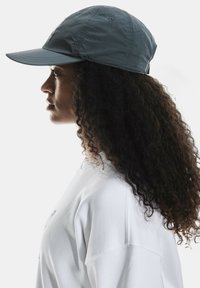 Cască gri cu margine curbă, fabricată dintr-un material ușor; are detalii de cusătură și orificii de ventilație. Purtați cu o cămașă albă.