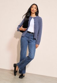 Moteris stovi prie sienos dėvėdama mėlyną megztinį su sagomis, baltą palaidinę, mėlynas džinsus, juodus batelius, rankoje laiko juodą delninę.