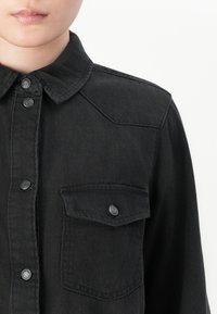 Person wearing a black denim shirt with metal snap buttons and a buttoned chest pocket visible on a white background.
