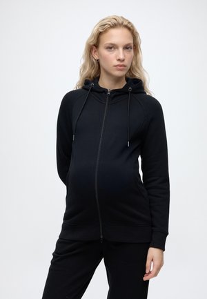 Zwangere vrouw met lang blond haar, een zwarte hoodie met rits en bijpassende broek dragend, staand tegen een effen lichte achtergrond.
