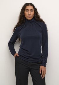 Navy-blaues langärmeliges Rollkragenshirt mit hohem Kragen, weichem Griff und gerafftem Detail am Ausschnitt, kombiniert mit schwarzen Hosen.