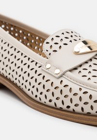Chaussure slip-on en cuir beige avec des détails perforés, un accent en métal doré sur la sangle et une semelle marron. Elle présente une texture lisse et une forme arrondie.